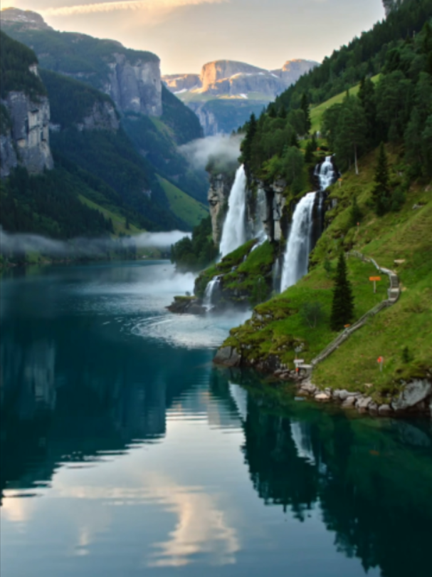 Geirangerfjord, Norway, Norway, with towering waterfalls cascading beside steep green cliffs reflected in calm fjord water beneath glowing morning light. #NatureLover #dreamplace #viral #fyp #geirangerfjord #norway